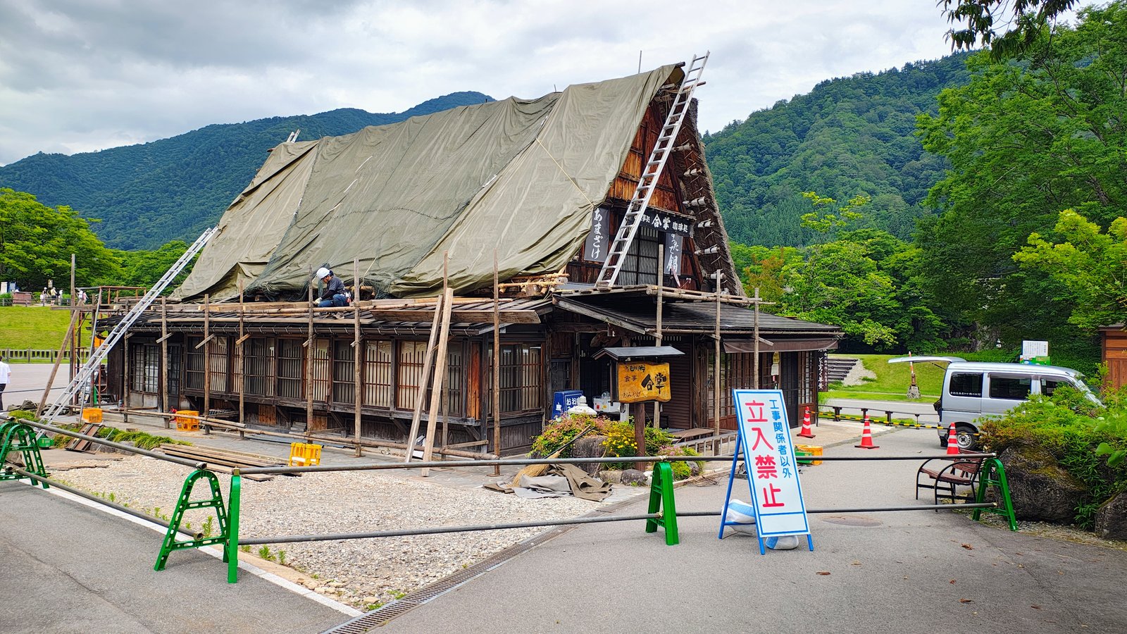 ช่างกำลังนั่งทำงานบนหลังคาหญ้าของบ้านกัสโชสึคุริ มีนั่งร้านไม้และบันไดพาดรอบอาคาร